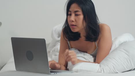 Junge-asiatische-Frau-mit-Laptop-Überprüfen-Social-Media-Gefühl-glücklich-lächeln,-während-auf-dem-Bett-liegen-duponieren,-nachdem-sie-morgens-im-Haus-aufwachen,-attraktive-thai-Mädchen-lächelnd-entspannen-im-Schlafzimmer-zu-Hause-Konzept.