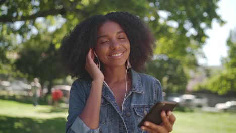 Eine-afrikanische-amerikanische-junge-Frau,-die-im-Park-steht-und-Musik-auf-Kopfhörern-auf-ihrem-Handy-hört---lächelnd-und-tanzende-Frau-im-Park