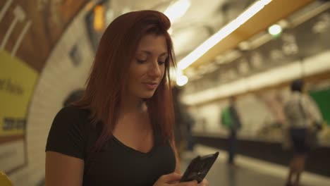 Atractiva-chica-pelirroja-con-pecas,-piercings-y-pelo-rojo-charlando-en-el-teléfono-inteligente-en-la-estación-de-metro,-durante-el-verano-soleado-en-París.-Fondo-subterráneo-borroso.-4K-UHD.