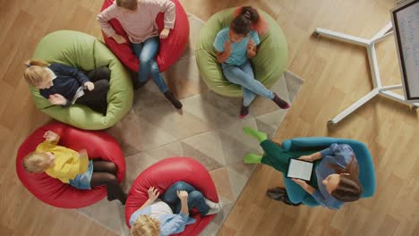 Elementary-School-Creativity-Classroom:-Children-Sitting-on-the-Bean-bags-while-Teacher-Explains-Lesson-to-them-and-Uses-Digital-Tablet-Computer,-They-Play-by-Passing-High-Five.-Top-View-Camera