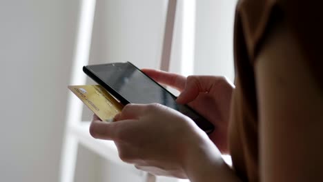 Close-up-hands-of-a-beautiful-Asian-caucasian-woman-using-smartphone-shopping-online-and-use-the-credit-card-payment-while-standing-beside-the-window-at-the-office-room.