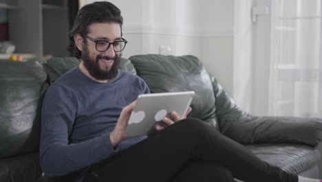 Young-Caucasian-man-with-black-hair-and-beard-wearing-eyeglasses-using-tablet-as-sitting-at-the-couch.-Intelligent-guy-using-social-media.-Internet-addiction,-facebook,-instagram.