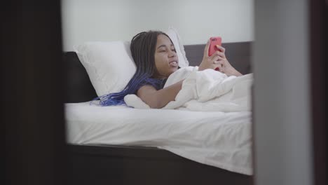 Portrait-of-thoughtful-girl-taking-smartphone,-typing-and-smiling.-African-American-teenager-resting-in-white-bed-at-home.-Social-media,-Internet-addiction,-lifestyle.