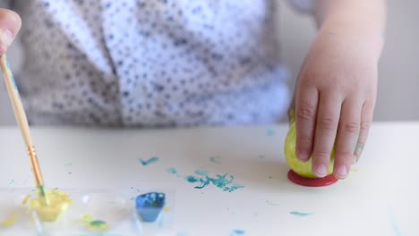 The-hands-of-a-litlle-child-paint-an-Easter-egg-in-yellow-color-on-a-stand.