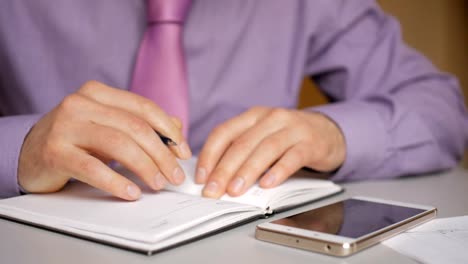 Businessman-in-a-purple-shirt-and-tie-makes-notes-in-a-notebook.-Man-writes-a-pen-in-daily.-A-person-makes-notes-in-organizer.-Laptop-and-smartphone-on-office-desk-in-office.-Closeup.