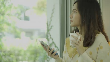 Nahaufnahme-von-4K-Aufnahmen-von-jungen-asiatischen-Frau-mit-Handy-und-Kaffee-trinken-in-der-Nähe-des-Fensters-am-Morgen.