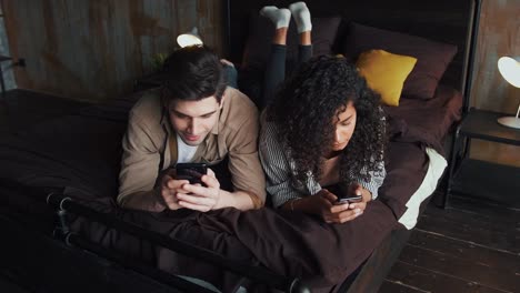 Spouses-in-casual-clothes-are-smiling,-using-their-smartphones.-They-lying-together-on-bed-in-room-with-modern-interior.-Stay-home.-Close-up