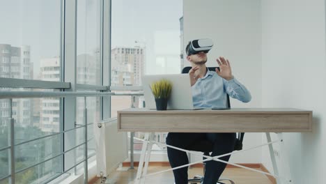 A-young-man-sitting-at-a-desk-in-the-office-uses-augmented-reality-glasses-to-work-on-business-projects-in-various-fields.