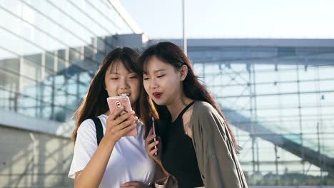 Vista-cercana-de-felices-y-encantadoras-chicas-asiáticas-en-ropa-de-moda-de-pie-frente-al-aeropuerto-y-mirando-el-teléfono