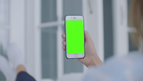 Sobre-la-toma-del-hombro-de-la-mujer-sosteniendo-un-teléfono-móvil-o-un-teléfono-inteligente-con-una-pantalla-verde-vertical-en-su-mano-en-casa-tumbada-en-el-sofá.-Chica-toque-en-la-pantalla-y-ver-el-contenido.