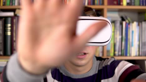 Junger-Mann-in-VR-Brille-in-der-Bibliothek.-Ein-Mann-mit-VR-Helm-auf-dem-Kopf-untersucht-und-berührt-die-virtuelle-Realität.-Im-Hintergrund-sind-Bücher-über-Bücherregale.-Buchbibliothek.