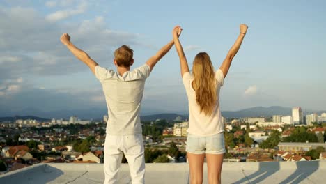 CLOSE-UP:-Two-lovers-standing-on-rooftop-above-the-city,-raising-hands-in-sky