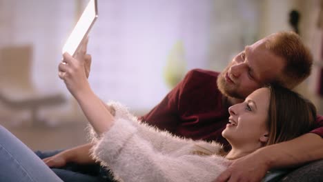 Oouple-joven-en-el-amor-en-el-sofá-haciendo-Inicio-selfie-con-tablet