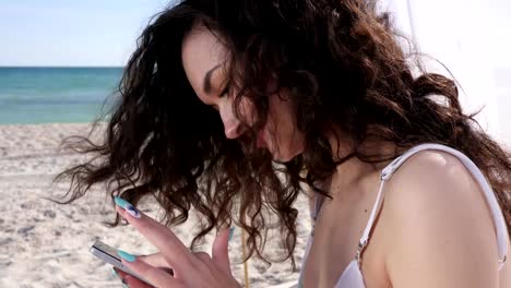 chica-hablar-en-las-redes-sociales-en-la-playa,-sonriendo-la-mujer-utiliza-móvil-en-la-costa-del-océano,-joven-Ver-perfil-en-el-teléfono-celular-durante-el-descanso-de-verano