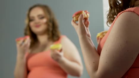 Chubby-female-enjoying-two-donuts-in-front-of-mirror,-admiring-her-body-shape