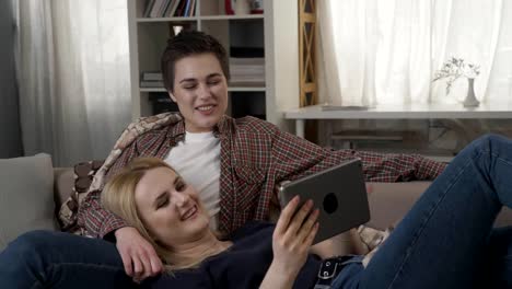 Lesbian-couple-is-resting-on-the-couch,-using-tablet-and-laughing,-young-family,-plaid,-cozy-60-fps