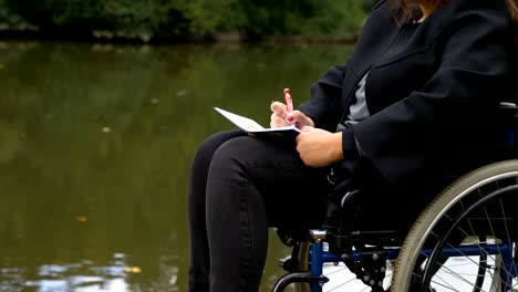 close-up-on-woman-on-wheelchairs-writes-a-love-letter-near-the-lake