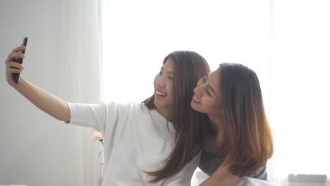 Slow-motion---Beautiful-young-asian-women-LGBT-lesbian-happy-couple-sitting-on-bed-hug-and-using-phone-taking-selfie-together-bedroom-at-home.-LGBT-lesbian-couple-together-indoors-concept.