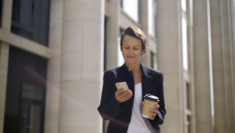 Sonrisa-medio-envejecido-a-mujer-con-pelo-corto-caminando-por-la-calle-con-taza-de-café-para-llevar-y-mensajes-en-su-celular-buscando-feliz-e-inspirado-de-texto