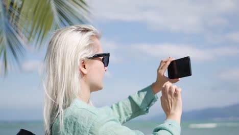 Junge-schöne-schlanke-Frau-mit-langen-blonden-Haaren-mit-Sonnenbrille-und-grünes-Hemd-stehen-in-der-Nähe-von-Palme-und-Fotografieren-von-Smartphone-auf-einen-blauen-Himmelshintergrund.-Mädchen-mit-Handy