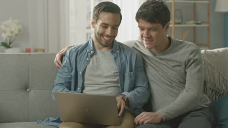 Adorable-Pareja-Gay-hombre-pasar-tiempo-en-casa.-Se-sientan-en-un-sofá-y-usar-la-Laptop.-Navegan-en-línea.-Pareja-pone-su-mano-alrededor-de-su-amante.-Habitación-tiene-un-Interior-moderno.