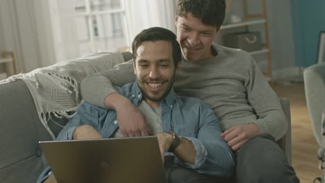 Linda-pareja-Gay-hombre-pasar-tiempo-en-casa.-Son-de-mentira-abajo-en-un-sofá-y-usar-la-Laptop.-Navegan-en-línea.-Mano-de-socio-es-alrededor-de-su-amante.-Sonreír-y-reír.-Habitación-tiene-un-Interior-moderno.