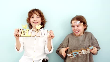 Studio-portrait-of-cute-school-children-with-wooden-signs.-Spring-holidays,-pastel-colors.-The-boy-is-wearing-a-white-shirt,-a-beige-dress-and-a-wreath-with-flowers