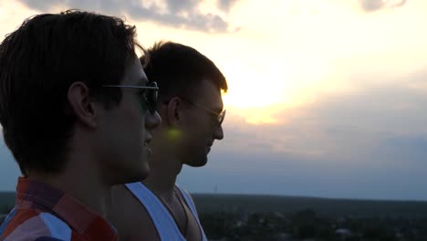 Profile-of-young-male-couple-in-sunglasses-standing-on-the-edge-of-rooftop-and-talking.-Handsome-gay-boys-relaxing-on-roof-of-high-rise-building-and-enjoying-beautiful-cityscape.-Close-up-Slow-motion