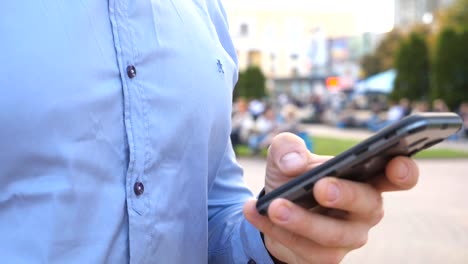 Nahe-Hand-von-jungen-Geschäftsmann-halten-und-berühren-Smartphone.-Unerkennbarer-Mann-steht-auf-der-städtischen-Straße-und-stöbert-Nachrichten-am-Telefon.-Guy,-der-Gadget-für-die-Arbeit-verwendet.-Langweilig-Dolly-erschossen