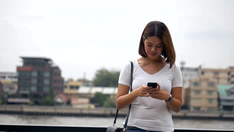 Schöne-junge-asiatische-Frau-mit-Smartphone-genießen-das-Lesen-von-sozialen-Medien-neben-Zaun-am-Fluss.