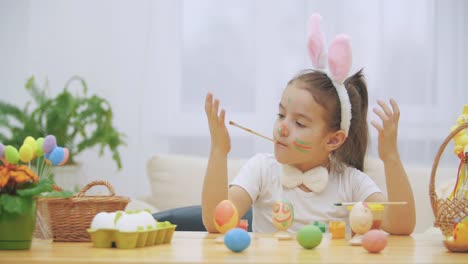 Nice,-cute-girl-is-having-fun-painting.-Adorable-girl-is-colorizing-and-her-hands,-one-and-another,-with-a-help-of-paint-brush-in-her-mouth.-Girl-with-beauty-spots-at-her-face-and-is-smiling-gently-with-cat's-whisker,-sitting-at-the-wooden-table-with-East