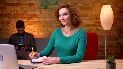 Closeup-shoot-of-adult-businesswoman-typing-on-the-laptop-taking-notes-looking-at-camera-and-smiling-indoors-in-the-office.-Female-and-male-employees-walking-on-the-background