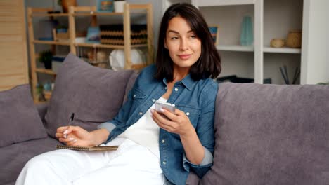 Slow-motion-of-happy-young-woman-writing-in-notebook-looking-at-smartphone