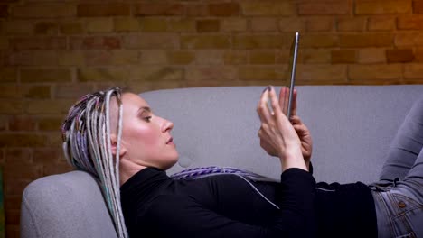 Closeup-shoot-of-young-attractive-caucasian-hipster-female-with-dreadlocks-using-the-tablet-turning-to-camera-and-happily-cheerfully-lying-on-the-couch-indoors-in-a-cozy-apartment