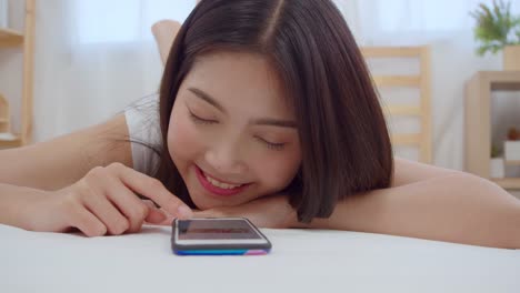 Slow-motion---Young-Asian-woman-using-smartphone-checking-social-media-feeling-happy-smiling-while-lying-on-bed-after-wake-up-in-the-morning,-Attractive-Japanese-girl-smiling-relax-in-bedroom-at-home.