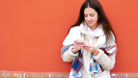 A-young-attractive-girl-stands-at-the-wall-of-a-building-and-pokes-a-finger-into-a-mobile-phone.