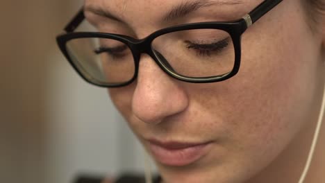 Close-up-of-attractive-cool-redhead-woman-with-glasses-reflection,-freckles-and-red-hair-watching-internet-video-on-smartphone-sitting-at-bus-stop-in-street,-during-sunny-summer-in-Paris.-High-angle.