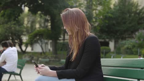 Business-woman-using-smartphone-Sitting-on-bench-in-park-in-Paris,-texting-with-mobile-telephone-outside,-typing-text-message-on-cell-phone.-Content,-Videos,-Red-hair.-Social-media-outdoor.-4K-UHD.