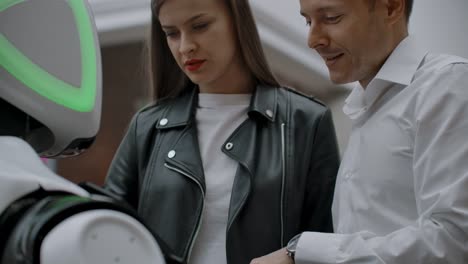 Un-hombre-y-una-mujer-en-el-centro-comercial-interactúan-con-un-consultor-de-robots-tocando-la-pantalla-y-sonriendo.-Cyborg-Android-ayuda-a-la-gente.