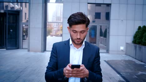 El-joven-hombre-de-negocios-utiliza-el-teléfono-inteligente-mientras-camina-por-la-calle-del-distrito-de-negocios-de-la-gran-ciudad.-Vestida-clásicamente.-Gran-edificio-de-oficinas-en-segundo-plano.-Mirando-con-éxito,-confiado.