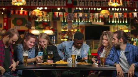 Grupo-de-amigos-de-estudiantes-de-bar-beben-cerveza-y-miran-las-pantallas-de-los-teléfonos-inteligentes-sonriendo