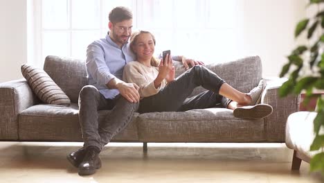 Pareja-feliz-riendo-sosteniendo-teléfono-hablando-relajantes-juntos-en-el-sofá