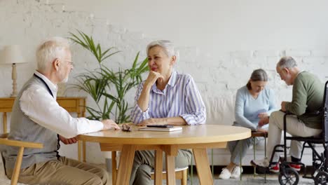 Tracking-shot-of-elderly-couple-sitting-at-table-and-enjoying-friendly-talk,-senior-woman-and-disabled-senior-man-in-wheelchair-playing-cards-in-background-in-common-room-of-nursing-home