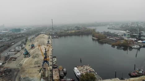 Luftaufnahme-der-industriellen-grauen-Stadt-mit-Nebel-und-Smog-bedeckt.-Güterzüge-und-Bahnhof-in-der-Nähe-von-Flussdocks-und-Frachthafenkranen