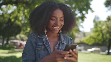 Lächelndes-Porträt-einer-afrikanischen-amerikanischen-jungen-Frau-mit-Kopfhörern-in-den-Ohren-genießen-chatten-und-nehmen-auf-Handy-im-Park-lächelnd