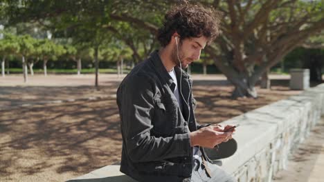 Retrato-de-un-joven-skater-joven-sonriente-con-auriculares-en-los-oídos-enviando-mensajes-en-el-teléfono-móvil-en-el-parque