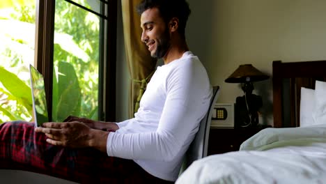 Hispanische-Jüngling-mit-Laptop-Computer-glücklich-lächelnden-Mann-im-Chat-Online,-über-große-Fenster-mit-Blick-auf-den-tropischen-Garten