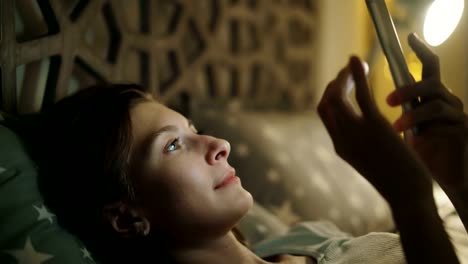 Close-up-of-young-smiling-woman-using-smartphone-lying-in-bed-at-home-at-night