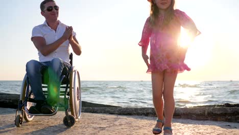 disabled-man-in-wheelchair-looks-at-jumping-child-in-backlight-near-sea-in-slow-motion-applauds-daughter