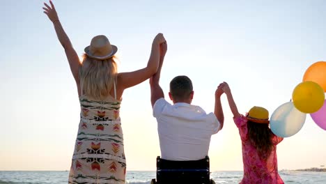 Invalid-with-wife-and-daughter-raise-their-hands-up-with-colorful-air-balloons-near-sea,-family-with-child